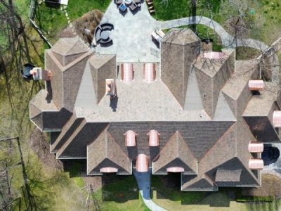 Aerial view of a large, multi-gabled house with a patterned roof, surrounded by trees, patio furniture, and green lawn.