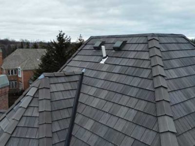 Gray metal shingle roof with two vent pipes and two roof vents, viewed from above on a cloudy day. Neighboring buildings and trees are visible in the background.