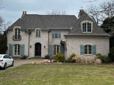 Two-story brick house with gray roof, blue shutters, and arched windows, surrounded by bare trees; a white car is parked in the driveway.