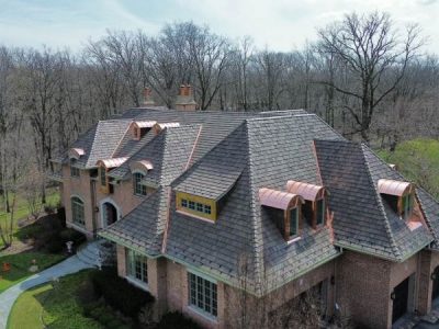 Large brick house with multiple gabled roofs, copper roof accents, and dormer windows, surrounded by leafless trees in a wooded area.