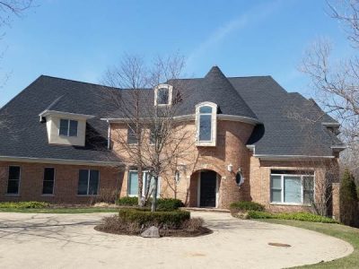 A large two-story brick house with black shingle roof, turret, arched entryway, and circular driveway bordered by leafless trees.