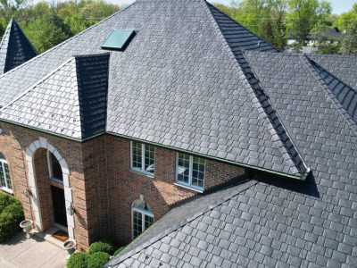Aerial view of a large brick house with a dark gray, shingled, multi-pitched roof and a skylight.