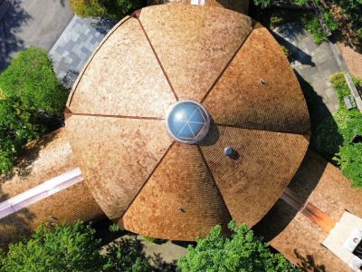Aerial view of a circular wooden building with a central glass dome, surrounded by trees and walkways.