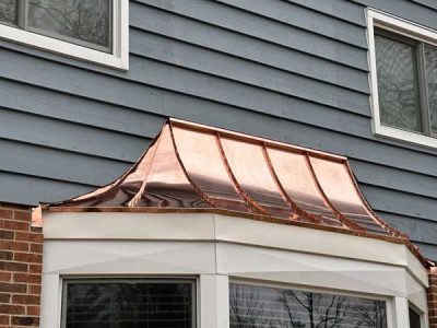 A copper roof awning with a curved design is installed above a bay window on the exterior of a house with blue siding and brick.